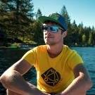 Florian Mauduit. Image shows a person dressed in a bright yellow shirt with a black graphic on it, wearing sunglasses and a dark colored baseball cap. Background is a body of water and evergreen trees.